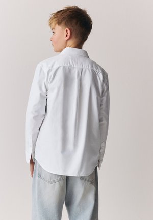 Junge, der seitlich schaut, trägt ein weißes Langarmshirt und hellblaue Jeans vor einem einfarbigen hellen Hintergrund.