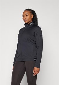 Zwarte ritssluitingjack van zacht materiaal, met een hoge kraag en zijzakken. Logo op de kraag en de onderste zoom. Gecombineerd met zwarte leggings.