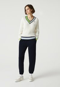 Biały sweter w warkocze z dekoltem w serek, w zielone i niebieskie paski, zestawiony z ciemnogranatowymi dresami i białymi sneakersami z elastycznymi mankietami.