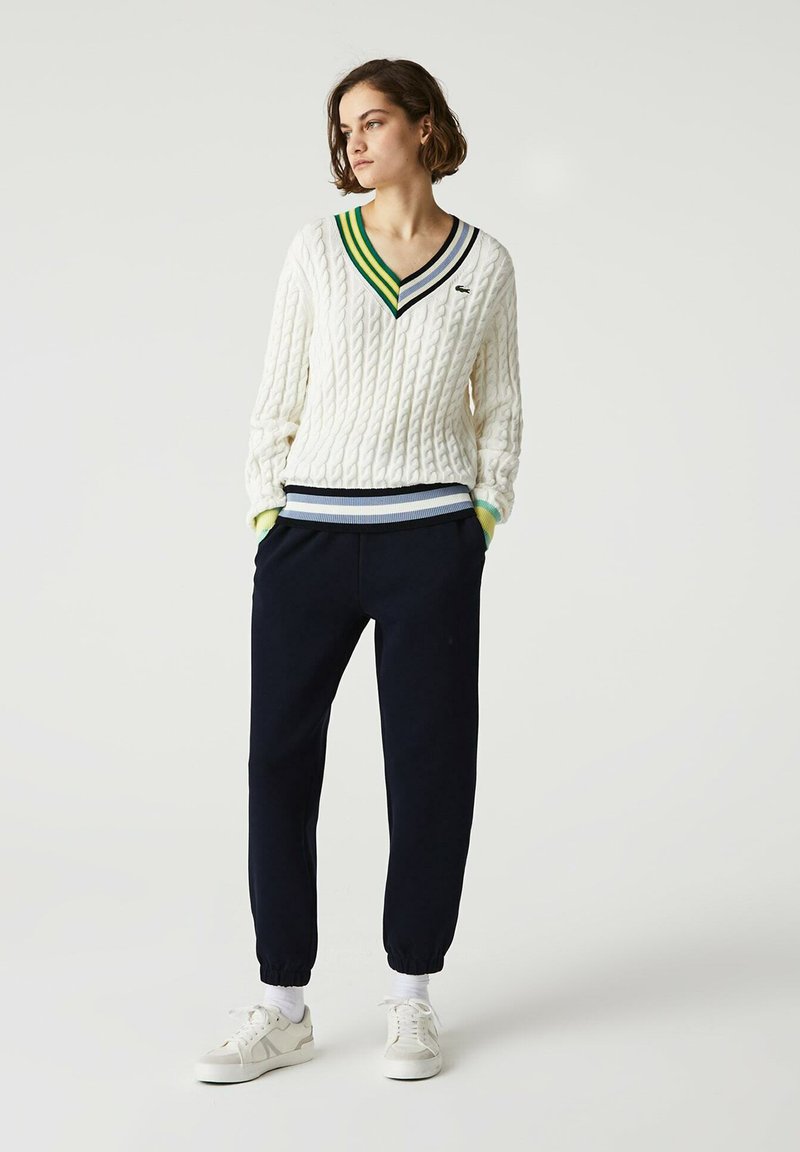 Biały sweter w warkocze z dekoltem w serek, w zielone i niebieskie paski, zestawiony z ciemnogranatowymi dresami i białymi sneakersami z elastycznymi mankietami.