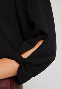 Blusa nera con tessuto testurizzato, maniche legate e aperture che rivelano la pelle. Design morbido e leggero.