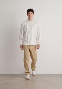 Biała koszula z długim rękawem z małym czarnym logo; beżowe chinosy; białe sneakersy z brązowymi podeszwami; luźny krój, gładka bawełniana tkanina.