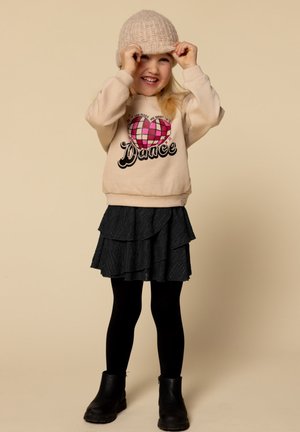 Jong kind dat glimlacht, een gebreide muts aanpast, een beige sweatshirt met een roze hart en de tekst "Dance" draagt, een zwarte rok, panty’s en laarzen.