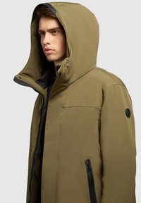 Olivengrøn isoleret jakke med aftagelig hætte, sort lynlås og sidelommer. Har glat stof og en sort logo-patch.