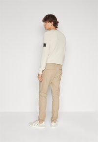 Pull crème avec une finition texturée, comportant une patch noire sur la manche, associé à un pantalon chino beige et des baskets blanches.