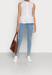 Haut peplum blanc sans manches avec des plis verticaux, devant à boutons, associé à un jean skinny bleu clair et un sac fourre-tout en cuir marron. Baskets blanches.