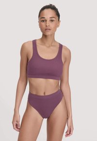 Vrouw met haar in een bun die een geribbelde paarse sportbeha en bijpassende hooggetailleerde onderbroek poseert tegen een effen lichte achtergrond.