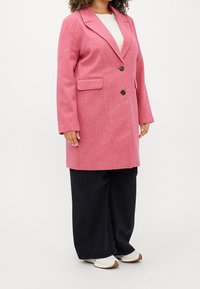 Abrigo de lana rosa con cuello muesca, bolsillos frontales y dos botones negros. Combinado con pantalones de pierna ancha negros y zapatillas blancas.