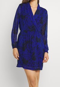 Blå blommig klänning med långa ärmar, omlottdesign och elastisk midja. Har svarta blommönster på ett genomskinligt tyg. Kort längd.