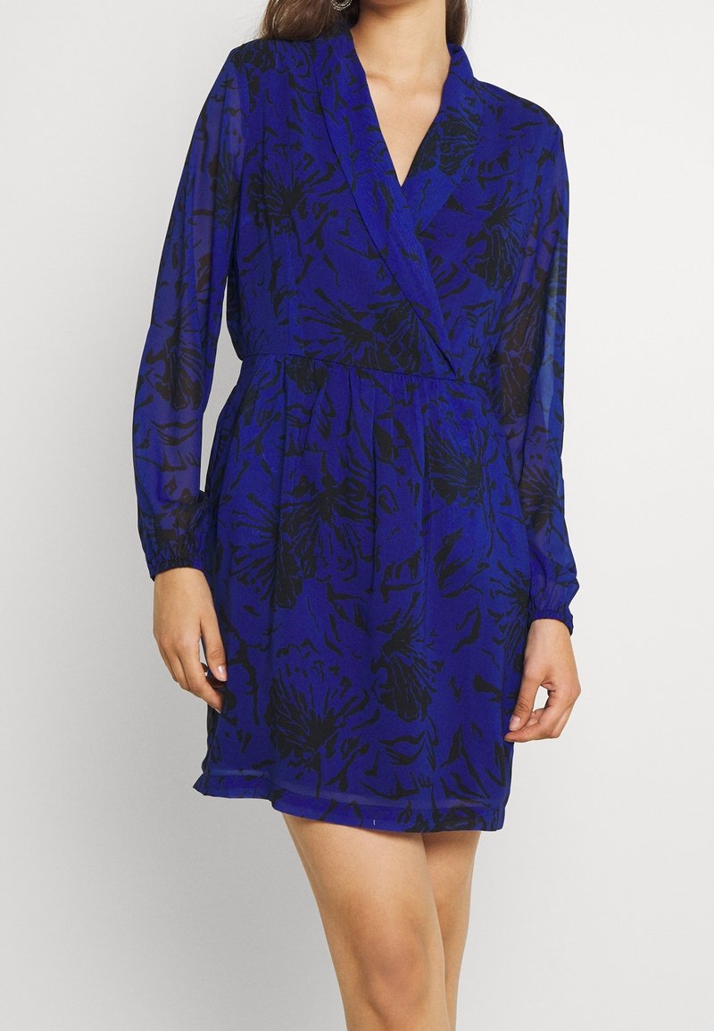 Blå blommig klänning med långa ärmar, omlottdesign och elastisk midja. Har svarta blommönster på ett genomskinligt tyg. Kort längd.