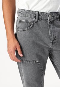 Pantalon en denim gris avec une finition texturée, présentant une fermeture à bouton sur le devant, des poches latérales et une étiquette de marque sur la ceinture.