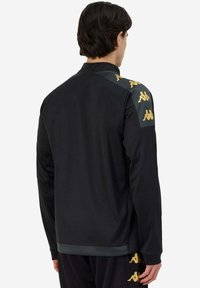 Chaqueta deportiva negra con cuello alto, mangas largas y logo de Kappa en dorado en los hombros. Hecha de material sintético suave.