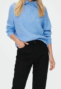 Suéter de punto azul claro con cuello con cremallera, con textura acanalada y mangas largas, combinado con pantalones negros de tiro alto.