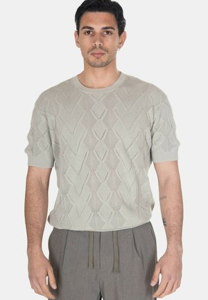 Uomo che indossa un maglione lavorato a maglia a maniche corte grigio chiaro con motivo a rombi e pantaloni grigi con coulisse, in piedi su sfondo bianco.