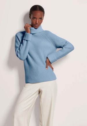 Vrouw poseert in een lichtblauwe coltrui en off-white wijde broek tegen een effen lichte achtergrond.