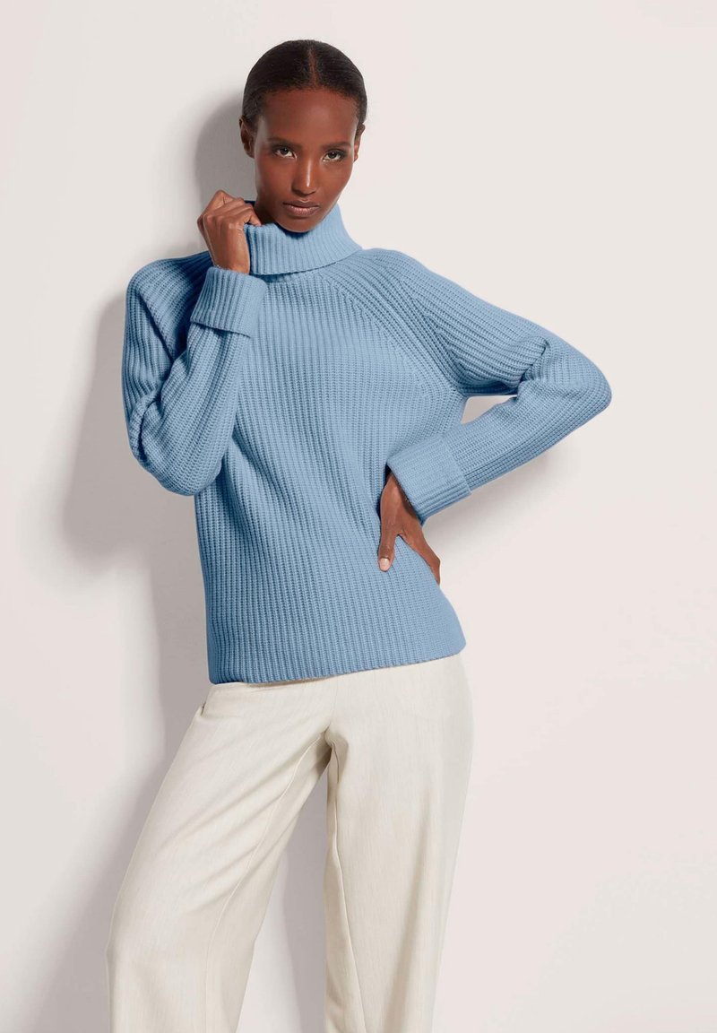 Frau posiert in einem hellblauen Rollkragenpullover und einer cremefarbenen, weiten Hose vor einem schlichten, hellen Hintergrund.