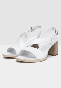 Sandales en cuir blanc avec un design entrecroisé, bout ouvert et bride de cheville. Talon bloc avec une apparence en bois texturé. Semelle plate.