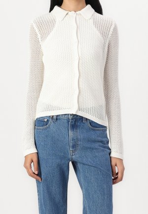 Vrouw draagt een witte mesh gebreide lange mouwen cardigan met knopen en blauwe high-waisted jeans, staande tegen een effen achtergrond.