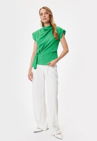 Top verde con scollatura drappeggiata, maniche corte e vita legata, abbinato a pantaloni bianchi a gamba larga e ballerine punta.