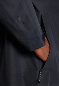 Manche d'une veste imperméable gris foncé présentant un logo de marque, une fermeture velcro et un accent à cordon au bas avec une texture lisse.