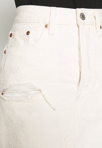 Bílá opotřebovaná džínová sukně s třepícími se okraji, měděnými nýtky a klasickým pětipocketovým designem. Texturovaná tkanina s viditelným švem.