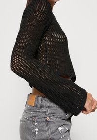 Pull noir court avec texture en tricot ajouré et manches longues évasées. Assorti à un jean gris avec des éclaboussures de peinture.