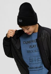Zwarte gewatteerde bomberjack, blauwe t-shirt met bedrukte tekst en een basketbalveld grafiek, zwarte gebreide beanie met logo.
