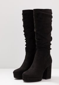 Botas negras de ante hasta la rodilla, con un diseño fruncido, punta cuadrada y tacón alto grueso. Textura suave con suela de plataforma.