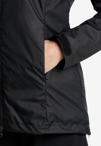 Veste extérieure noire avec une texture lisse, dotée d'une poche latérale et d'une fermeture à velcro au niveau des poignets. La fermeture éclair est bien visible à l'avant.
