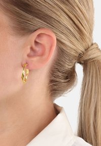 Blondine met haar opgestoken, die een gedraaid gouden oorring in haar linkeroor en een witte overhemd draagt.