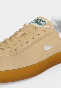 Бежова велурена маратонка Lacoste с гумена подметка, бежови връзки, бял вътрешен подплата и бяло лого на крокодил отстрани с текст "EST. 1933".