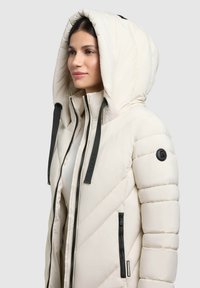 Giacca imbottita beige chiaro con un ampio cappuccio, design a imbottitura diagonale, chiusura frontale con zip e tasche, e zip e cordini a contrasto di colore scuro.