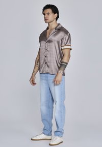 Camisa brillante de bronce de manga corta con botones en el frente, puños con acentos dorados, combinada con jeans de color azul claro y zapatillas blancas. Fondo liso.