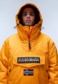 Žlutá zateplená bunda s vysokým límcem, přední klokankovou kapsou, značkovou nášivkou a vlajkovým emblémem. Obsahuje stahovací šňůrky na kapuci.