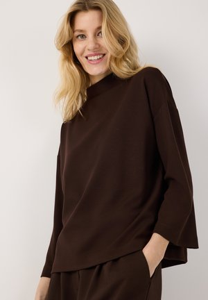Blonde Frau, die ein lockeres dunkelbraunes Langarmshirt und passende Hosen trägt, lächelnd mit einer Hand in der Tasche vor schlichtem Hintergrund.