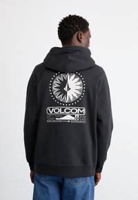 Fekete kapucnis pulóver, állítható zsinórral, a hátsó részén nagy fehér grafikával, amely egy iránytűt és szöveges részleteket ábrázol.