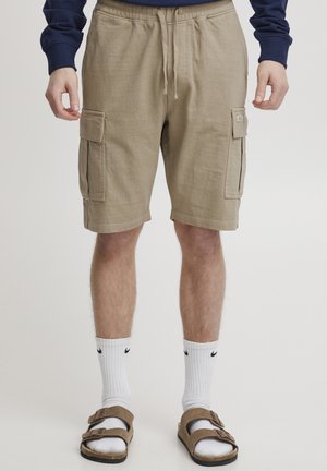 Helle braune Cargo-Shorts mit einem elastischen Bund, zwei seitlichen Taschen und einer weichen Textur, kombiniert mit weißen Socken und braunen Sandalen.