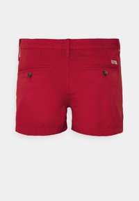Červené bavlněné šortky s hladkým povrchem, vybavené dvěma zadními kapsami s knoflíky a malou značkovou cedulkou na pravé straně.