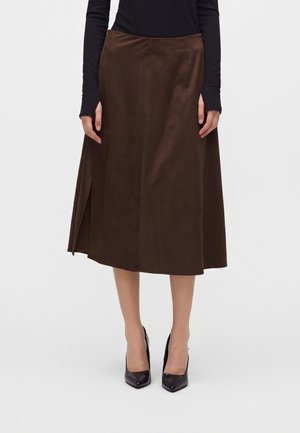 Femme portant une jupe midi marron avec une fente latérale, un haut noir à manches longues et des talons hauts noirs pointus, debout sur fond blanc.