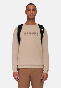 Felpa beige con scollatura rotonda e testo in contrasto marrone "MAMMUT SINCE 1862". Tessuto liscio, maniche lunghe, indossata con uno zaino nero.