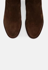 Botas de ante marrón hasta el tobillo con un acabado suave, puntera redondeada y detalles de costura sutiles, que presentan una silueta clásica y una textura suave.