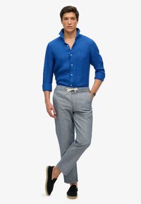 Blauwe linnen shirt met een knoopjeskraag, gecombineerd met grijze gestreepte elastische broeken, en zwarte instappers met een tan kleurige zool.