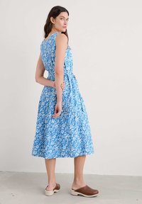 Rochie albastră cu un model floral alb, design fără mâneci, corp strâmt și fustă largă; asortată cu saboți maro.