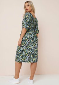 Schwarzes Blumenkleid mit kurzen Ärmeln und tailliertem Schnitt. Bunte Muster aus gelben und blauen Blumen, kombiniert mit weißen Sneakers.