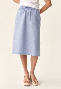 Donna che indossa una gonna in lino azzurro chiaro alla lunghezza del ginocchio, con vita regolabile e sandali platform bianchi, in piedi davanti a uno sfondo beige.