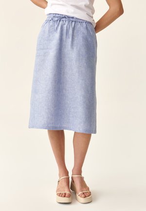 Femme portant une jupe en lin bleu clair jusqu'au genou avec une taille à cordon et des sandales compensées blanches, se tenant contre un fond beige.