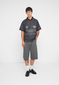 Svartmønstret kortermet skjorte med polo-krage, kombinert med grå kne-lange shorts. Modellen har på seg svarte sko og hvite sokker.