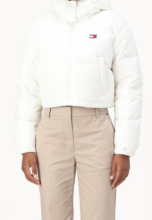 Personne portant une doudoune blanche courte avec un logo rouge, blanc et bleu marine sur la poitrine et un pantalon beige taille haute.