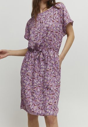 Kvinde iført en knælængde, lilla kjole med korte ærmer, blomsterprint og snøre i taljen, stående med den ene hånd i lommen.