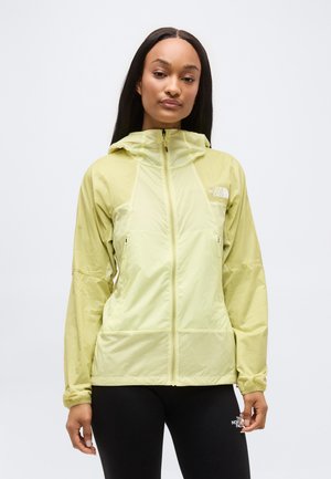 Femeie purtând o jachetă ușoară verde deschis cu fermoar și glugă și colanți negri, ambele branduite cu logo-urile The North Face.